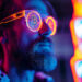 A man with a beard wearing glowing neon sunglasses with the Bitcoin logo on the lenses is illuminated by vibrant pink, blue, and orange lights in a futuristic setting. | Crypto.hu.net